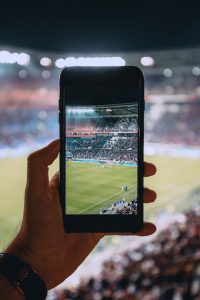 Ein Handy in der Hand nimmt ein Fußballspiel im Stadion auf.